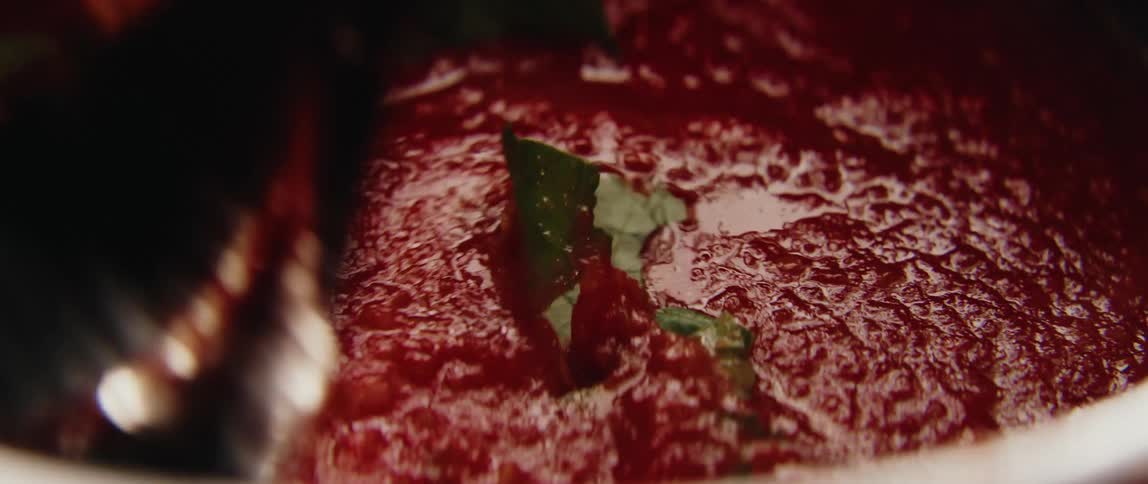 A person adds fresh basil leaves to a thick bubbling tomato sauce and stirs the mixture thoroughly with a wooden spoon in a white pot