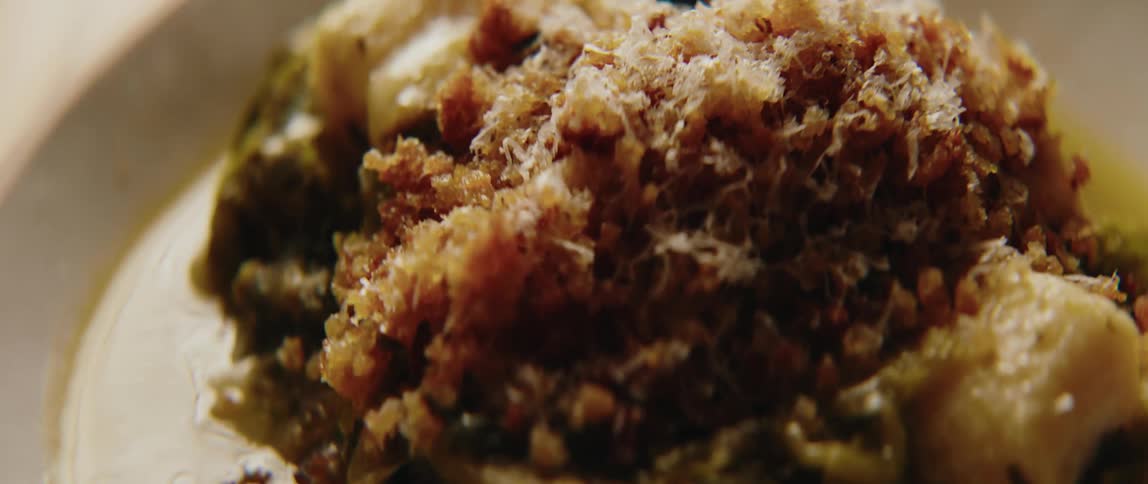 A close view shows a prepared dish of green vegetables topped with golden brown breadcrumbs and grated cheese in a white bowl