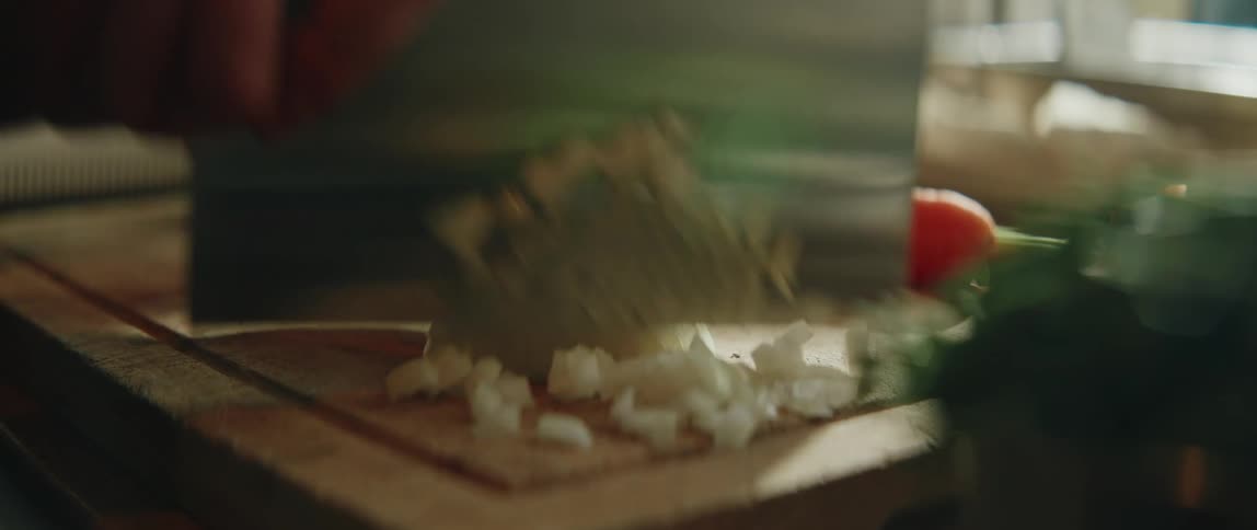 Onion being chopped on a wooden cutting board with a wideblade knife and vegetables in the background