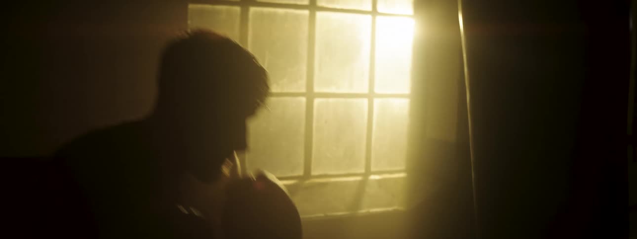 A shirtless man in silhouette intensely strikes a heavy punching bag in a dimly lit gym while golden sunlight streams through a gridded window illuminating around him as he trains