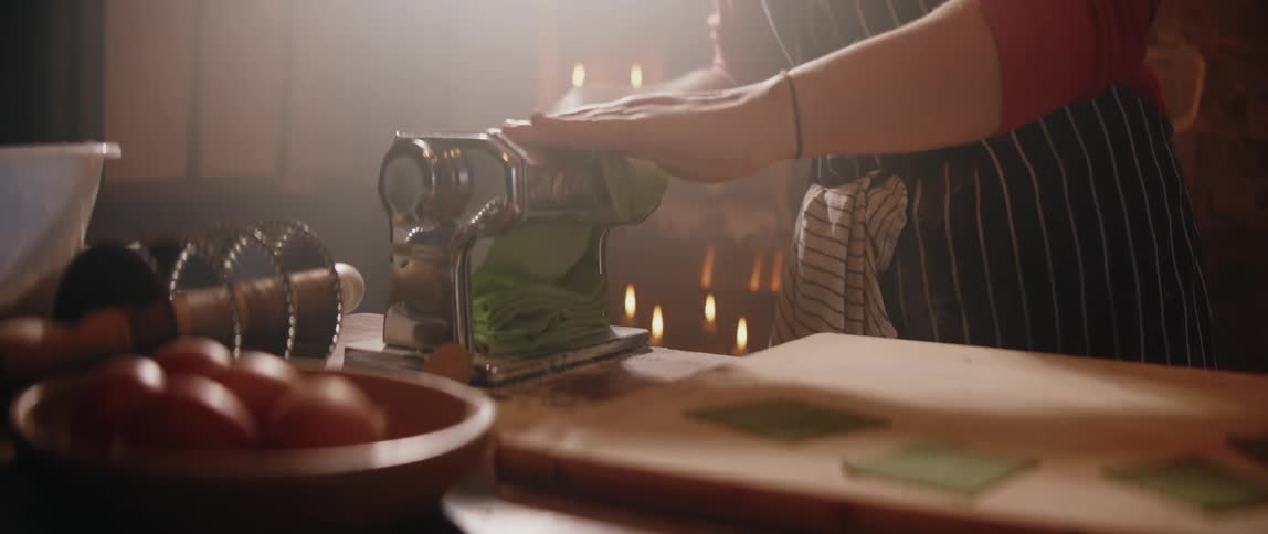 Person using a manual pasta machine to roll a strip of green dough on a wooden countertop