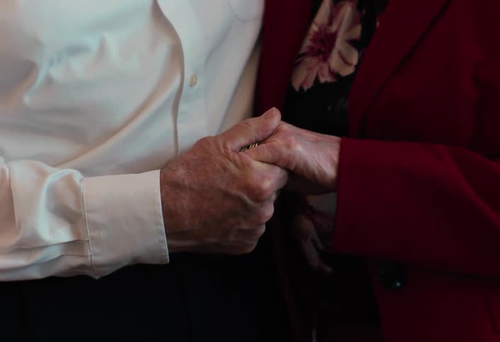Elderly couple standing together with his arm around her and they hold hands