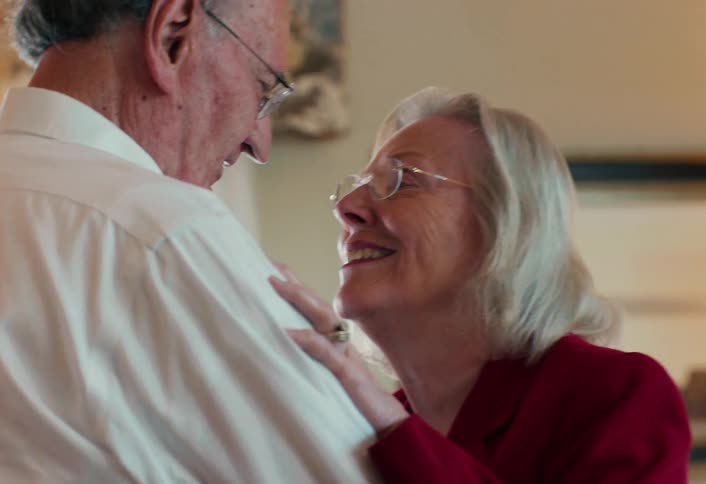 A close view shows an elderly woman with glasses and a red jacket smiling warmly at an elderly man in a white shirt while resting her hand on his chest