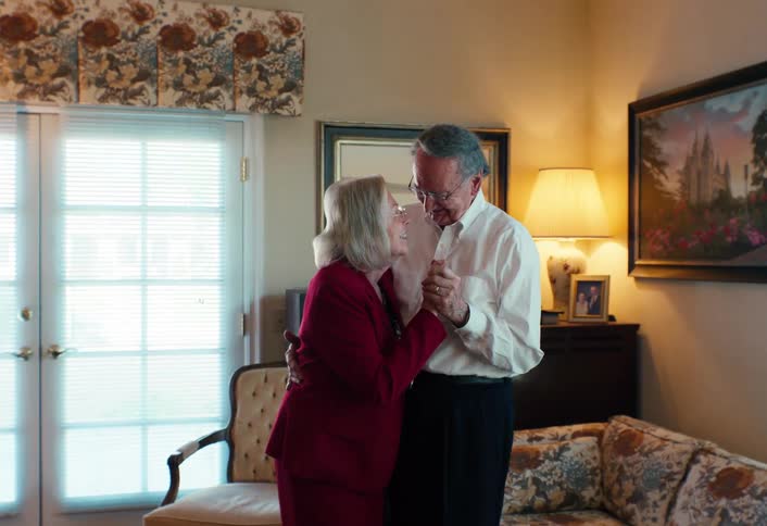 Elderly couple holding hands in a welllit living room with furniture framed artwork and a lamp nearby