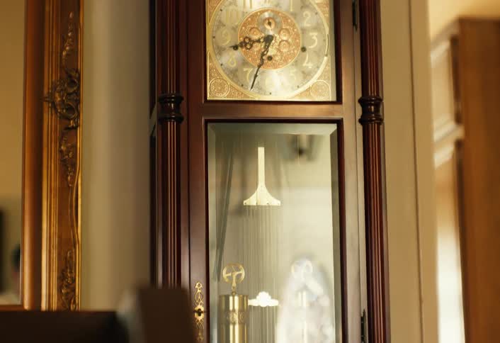 A dark wood grandfather clock stands against a wall next to a gold ornate mirror as its pendulum swings steadily behind glass and the internal brass weights remain visible below the decorative clock face