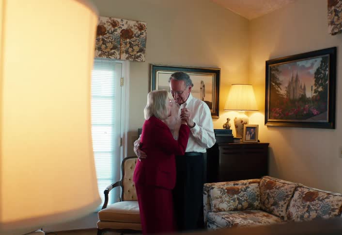 An older couple dances together in a warmly lit living room framed by furniture and soft background lighting