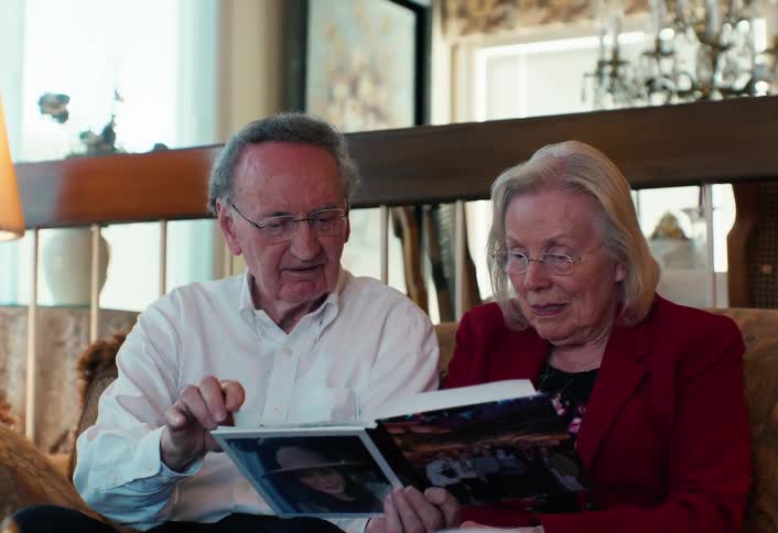 An elderly man and woman sit closely on a couch looking through a physical photo album together and pointing at the pictures while talking in a brightly lit living room