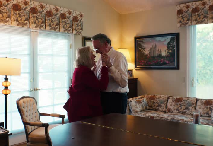 An elderly man in a white dress shirt and a woman in a red suit dance slowly together in a sunlit living room featuring a grand piano a floral couch and a large painting on the wall