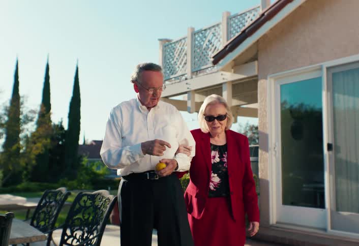 Elderly couple walking in a residential outdoor area with the man holding fruit and the woman wearing sunglasses beside him