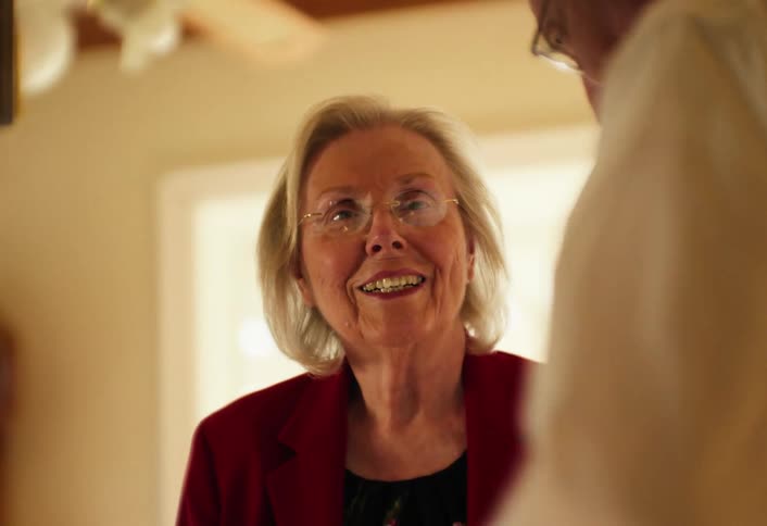 An elderly woman in a red jacket smiles warmly as she greets someone then turns and looks down