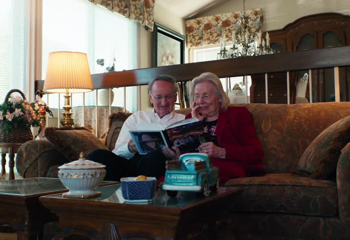 Elderly couple sitting on a couch reading a magazine in a living room with a coffee table and lamp