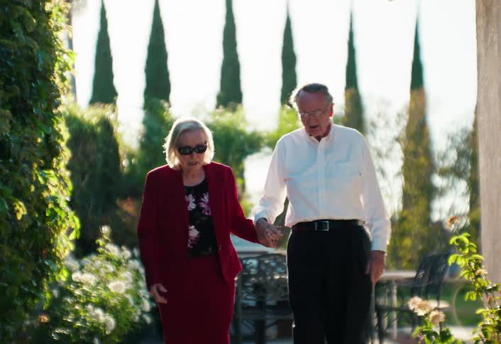 Elderly couple walking hand in hand through a residential garden with tall trees and outdoor furniture in the background
