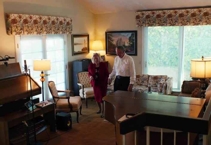 An elderly couple holding hands and smiling walk together through their cozy well lit living room past a piano and an organ