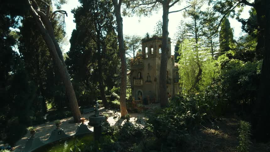 A stone tower with gothic arches stands nestled within a lush garden of tall pine trees and dense greenery as sunlight filters through the canopy near a small pond and fountain