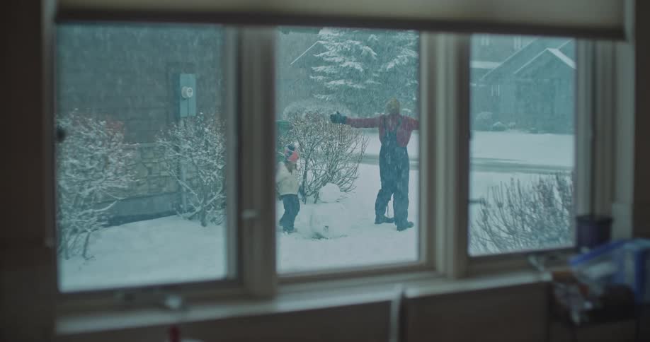 Through a kitchen window a father in red and his daughter are in their snowy backyard while thick flakes fall and the man raises his arms in a playful gesture near a residential street