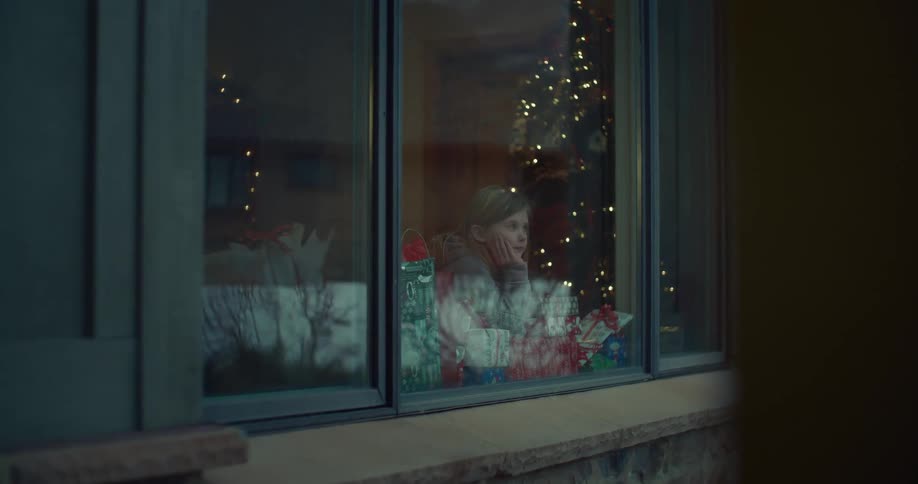 A young girl rests her chin on her hand and looks out a window surrounded by wrapped gifts and a glowing christmas tree reflected in the glass