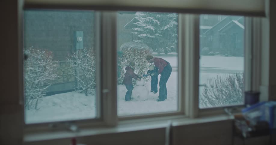 Through a window a father and his two young children work together to build a snowman in their snowy front yard while light snow falls across the suburban neighborhood