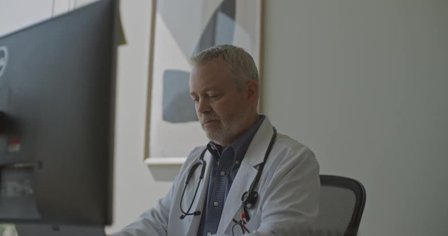 A senior male doctor in a white lab coat and stethoscope sits at a desk in a brightly lit office looking at a monitor and typing on a laptop