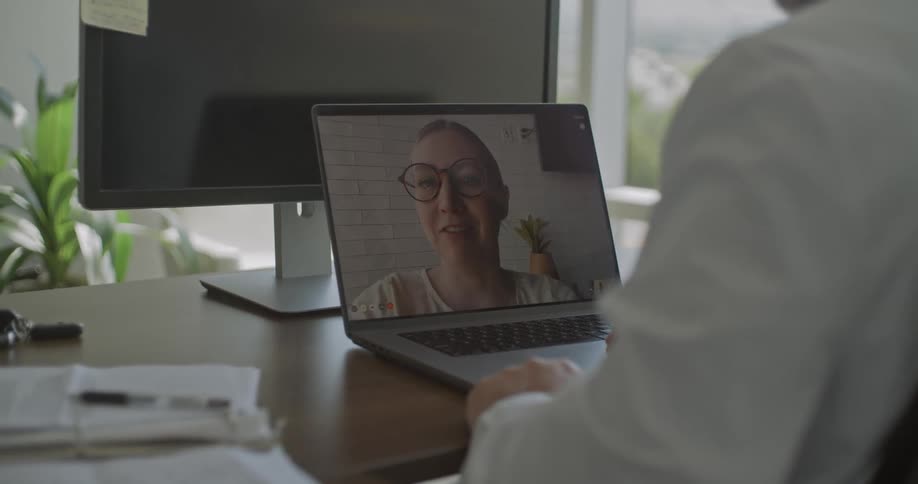 A person sits at a desk attending a video call on a laptop with a woman speaking on screen in a bright office setting