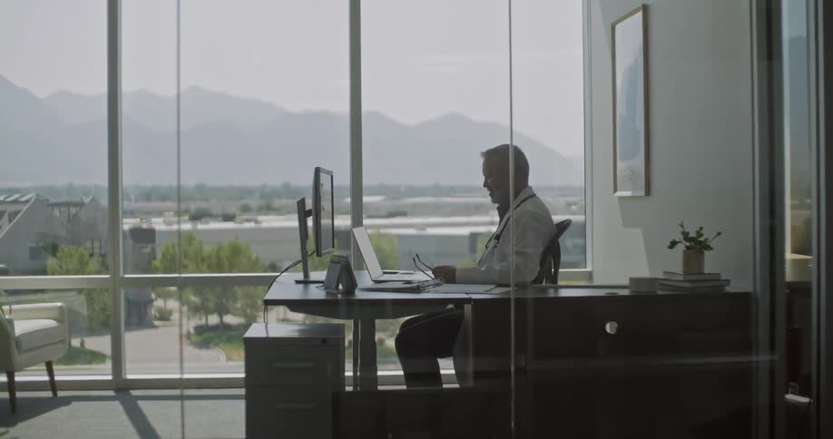 A man with lab coat seated in an office chair waving his hand to a laptop