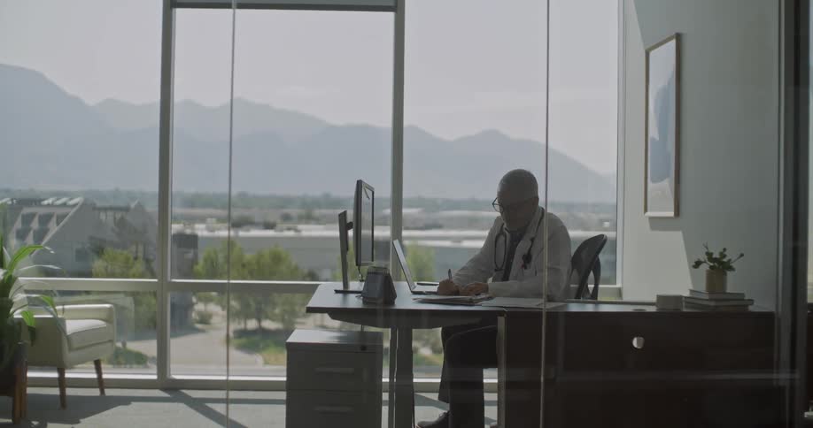Doctor sitting at a desk in his office with a laptop computer and monitor as he writes in a notebook