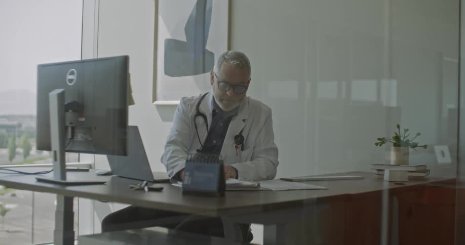 Doctor seated at a desk in a modern office writing on documents with a computer and work materials around