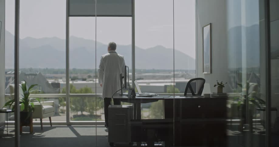 A male doctor sits at his desk in a glasswalled office closes his laptop stands up and walks toward a large window to look out at the mountains