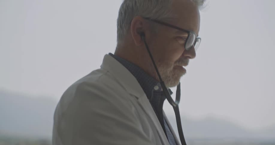 A grayhaired male doctor wearing a white lab coat puts on his stethoscope and places the chest piece on a young girls chest to listen to her heart while they stand in front of a bright window