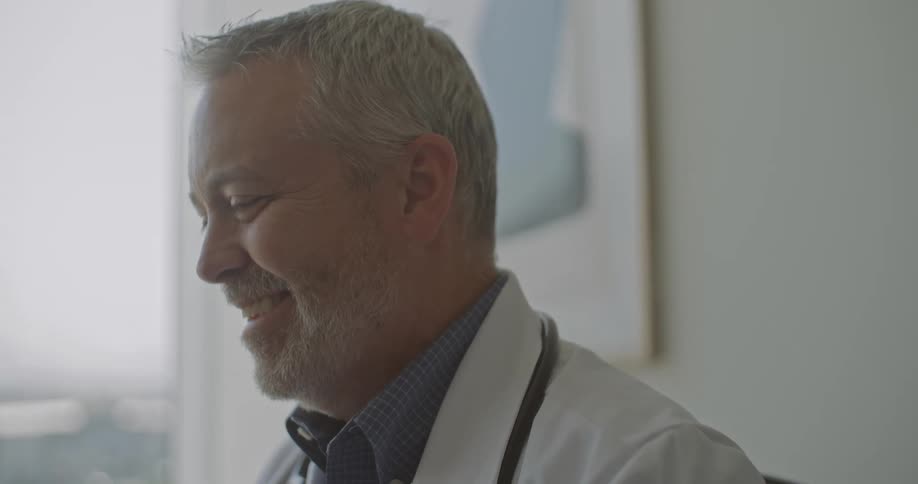 A middleaged male doctor with a grey beard and a stethoscope around his neck looks down and smiles warmly while sitting in a bright medical office