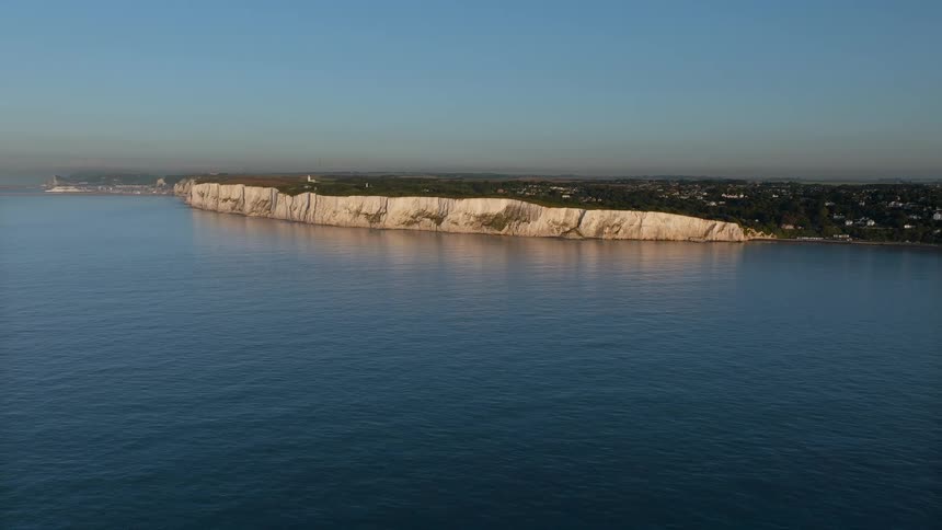 An aerial shot pans across cliffs as they meet the blue waters of the sea under a clear sky showing a small town and a lighthouse perched atop the grassy plateau during a bright calm morning