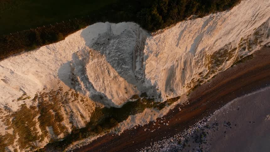 An aerial view shows sunlit white chalk cliffs rising sharply above a narrow shoreline and dark water below