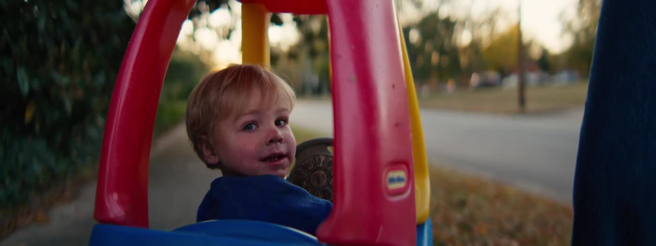 A young blond boy sits in a colorful plastic toy car on a suburban sidewalk glancing back over his shoulder with a joyful expression as the warm glow of the setting sun illuminates the leafy neighborhood behind him