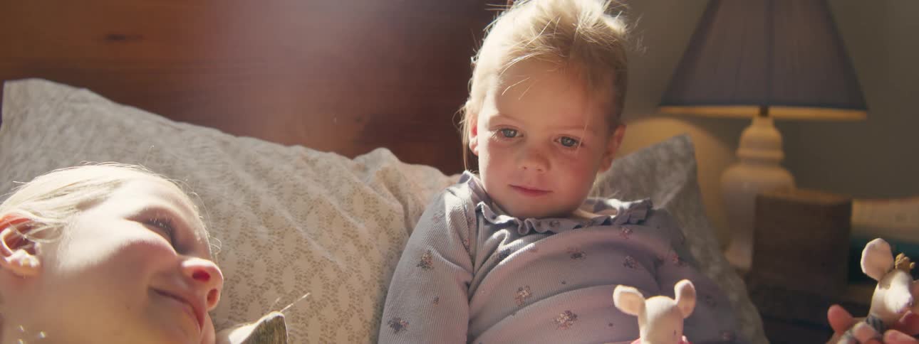 Children lying on a bed in a sunlit room holding toys and interacting with each other