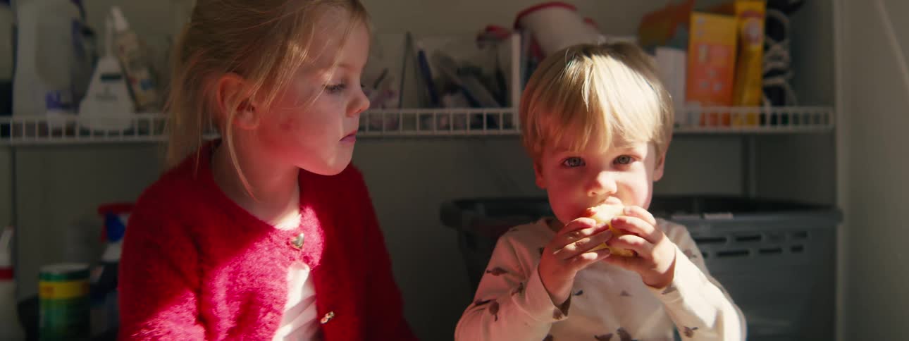 A young girl in a red cardigan watches as a toddler boy sitting beside her takes a large bite of a red apple in a sunlit pantry or kitchen area filled with storage bins and shelves