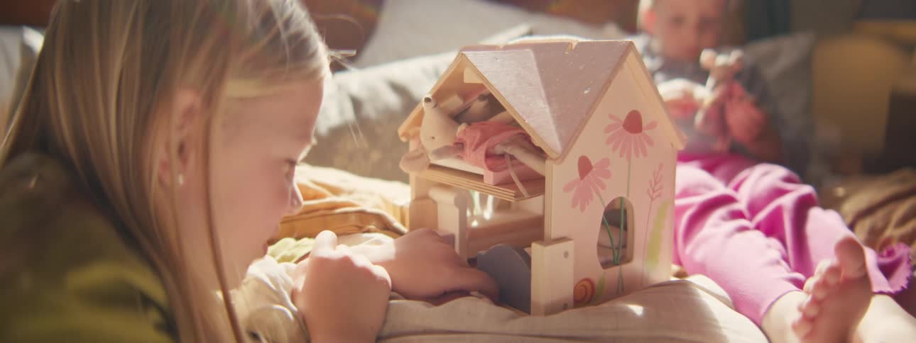 Little girl playing with her dollhouse as her brother plays with toys in the distance
