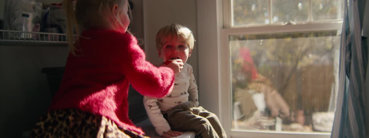 Child offering an apple to another child sitting near a window with natural light illuminating the indoor space