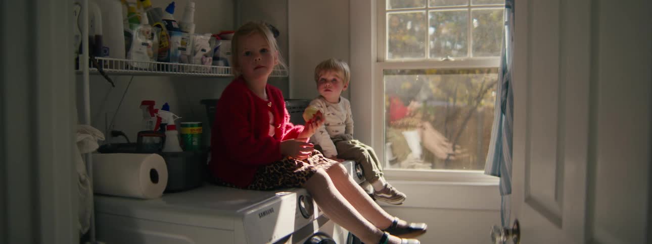 Little girl sitting on the washing machine in the laundry room eating an apple with her little brother looking around