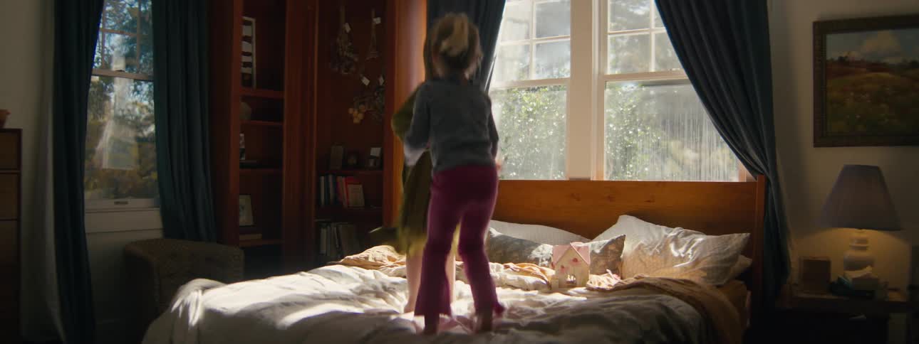 Two young girls joyfully jump and play on a bed together in a sunlit bedroom