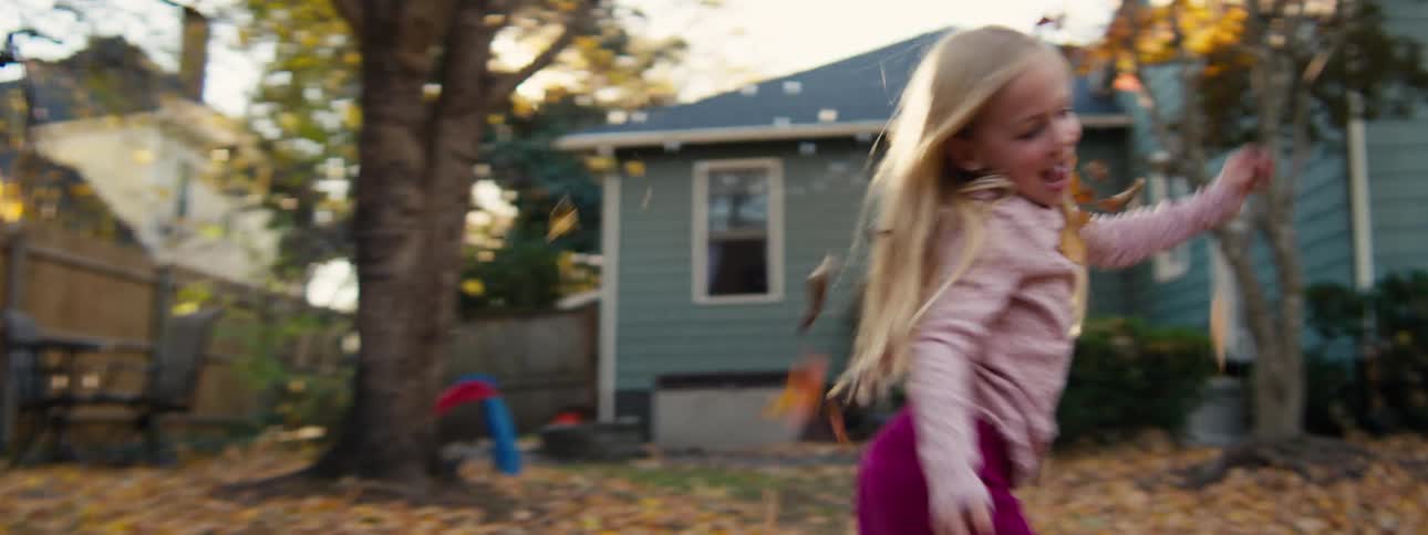 A young girl with long blonde hair and a pink sweater laughs as she twirls and plays in a backyard filled with falling autumn leaves under bright sunlight
