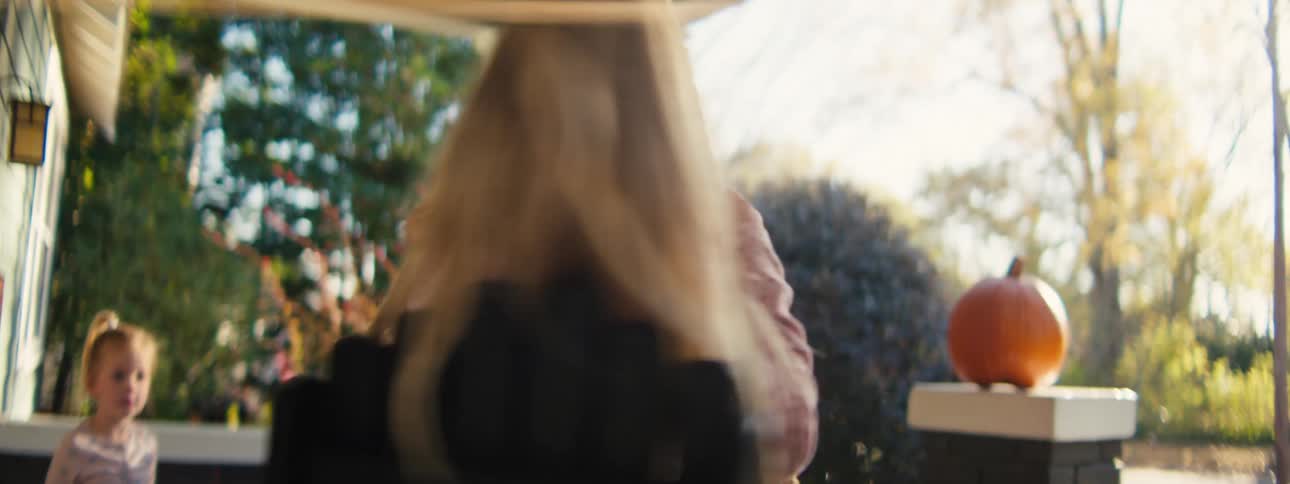 A young girl with long blonde hair and a pink sweater smiles and laughs while swinging on a rope swing on a porch