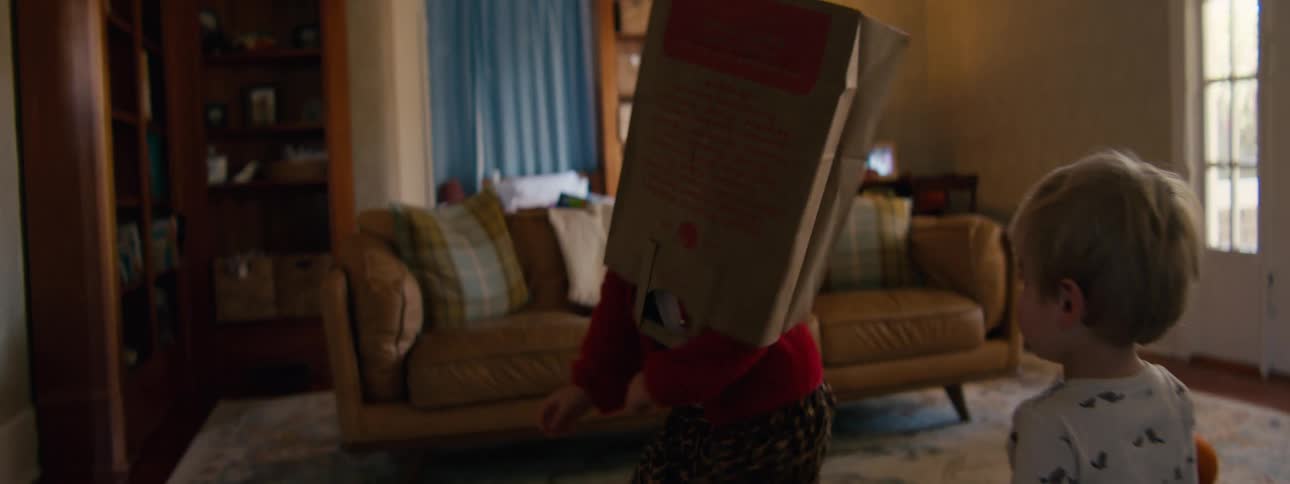 Little girl with a paper bag over her head playing in the living room as her little brother watches