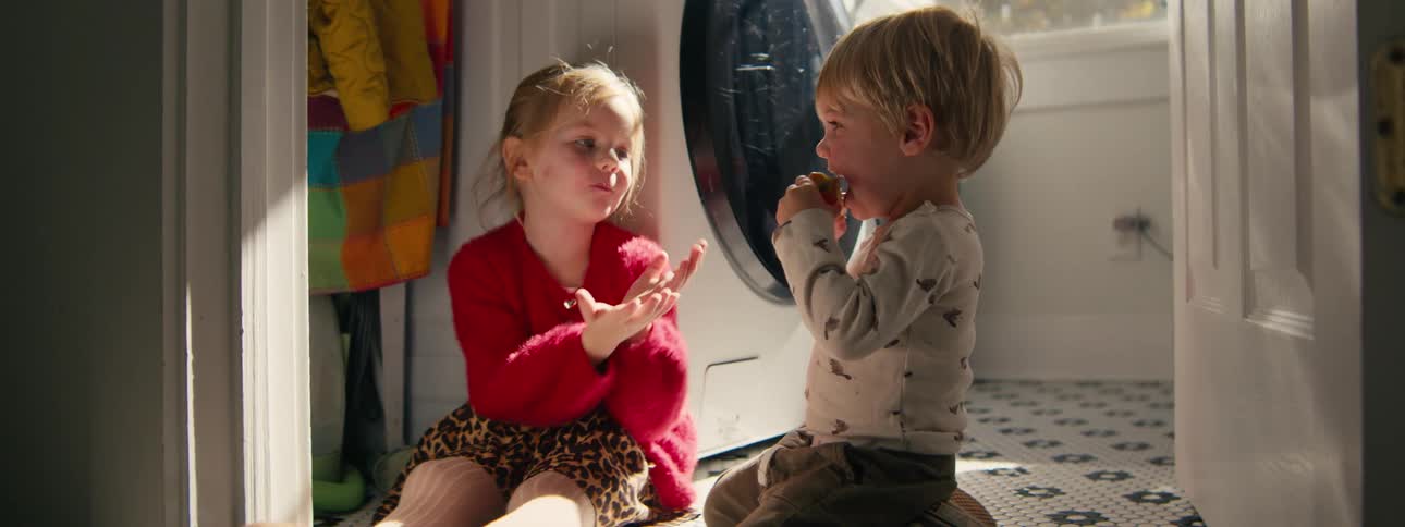 Two children sit on the floor of a domestic space eating fruit under natural light