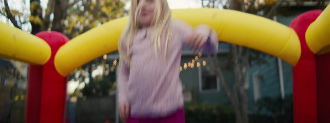 A young girl with long blonde hair and a purple sweater jumps excitedly inside a colorful yellow and red inflatable bounce house in a backyard