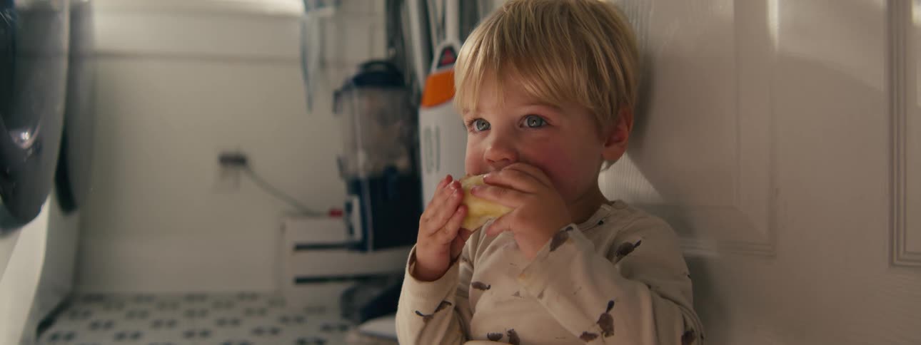 A young blond boy leans against a white wall in a kitchen happily munching on a slice of fresh fruit while glancing around the room with wide curious eyes