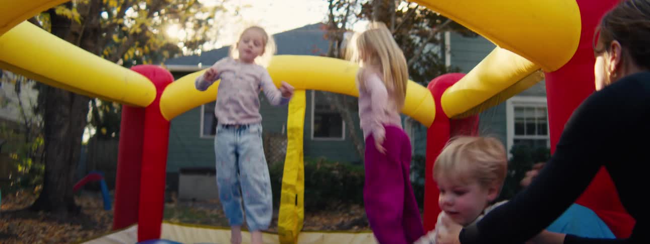 Two young girls and a toddler jump and play inside a colorful inflatable bouncy castle in a residential backyard during a sunny autumn afternoon while trees with yellow leaves and a house stand in the background