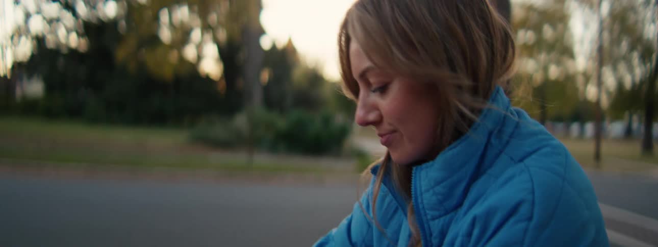 A woman wearing a bright blue puffer jacket looks down with a gentle smile while sitting outdoors on a sidewalk as the soft warm light of late afternoon illuminates the park trees in the background