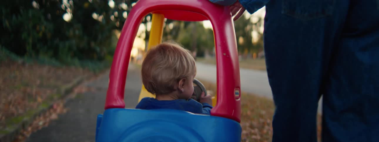 A mother pushing her son in a colorful toy car along a quiet path outdoors
