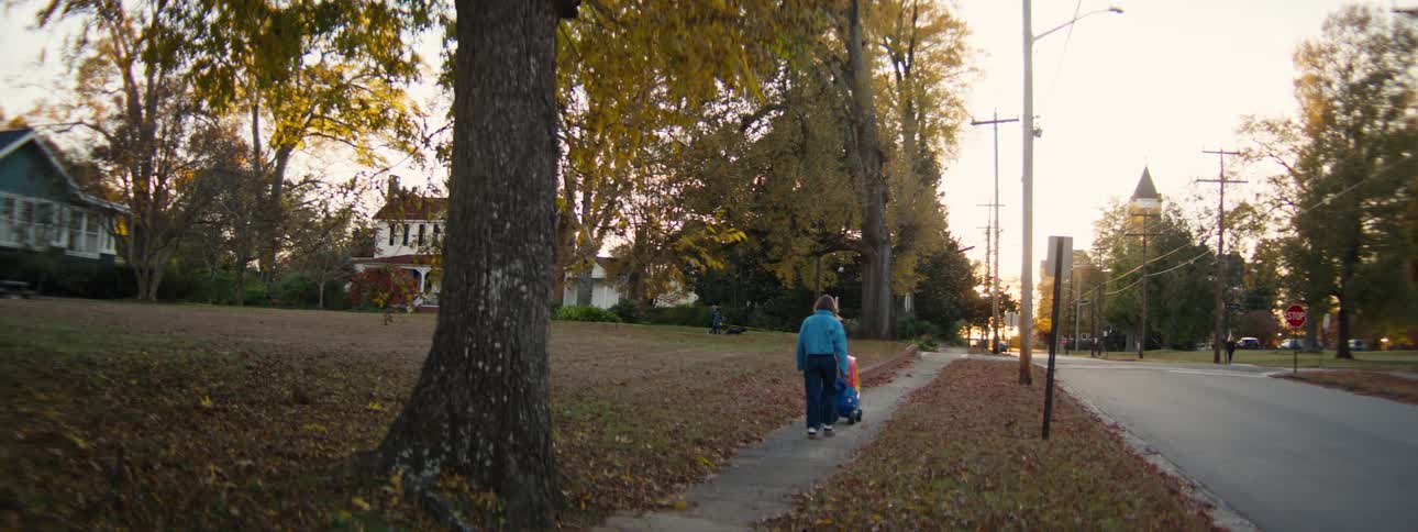 Mother follows as her child uses a toy vehicle on the sidewalk near a park on an autumn day