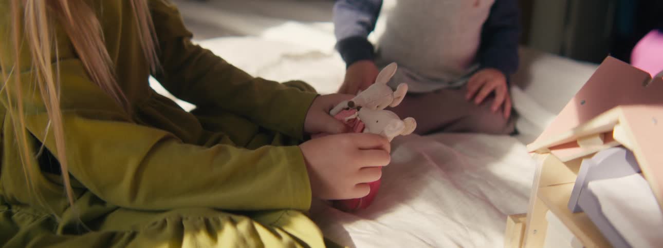 A young girl with long blonde hair sits indoors and plays with two small white mouse figurines she gently adjusts the fabric clothing on the toys while interacting with them on a soft surface