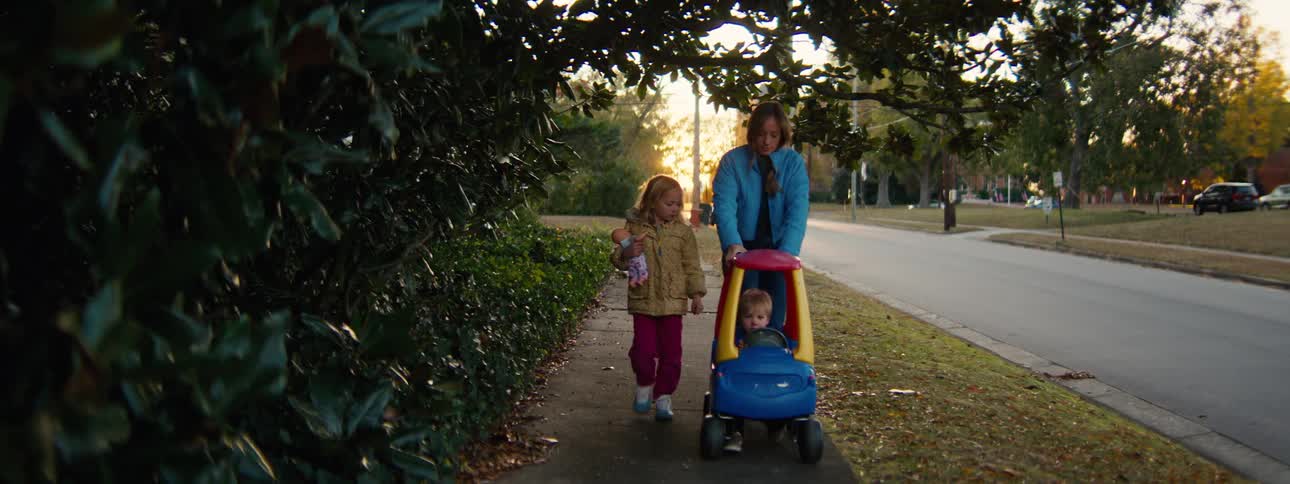 A medium shot shows a woman in a blue jacket pushing a toddler in a colorful toy car along a suburban sidewalk at sunset while a young girl with a doll walks beside them and another child follows behind on a bicycle